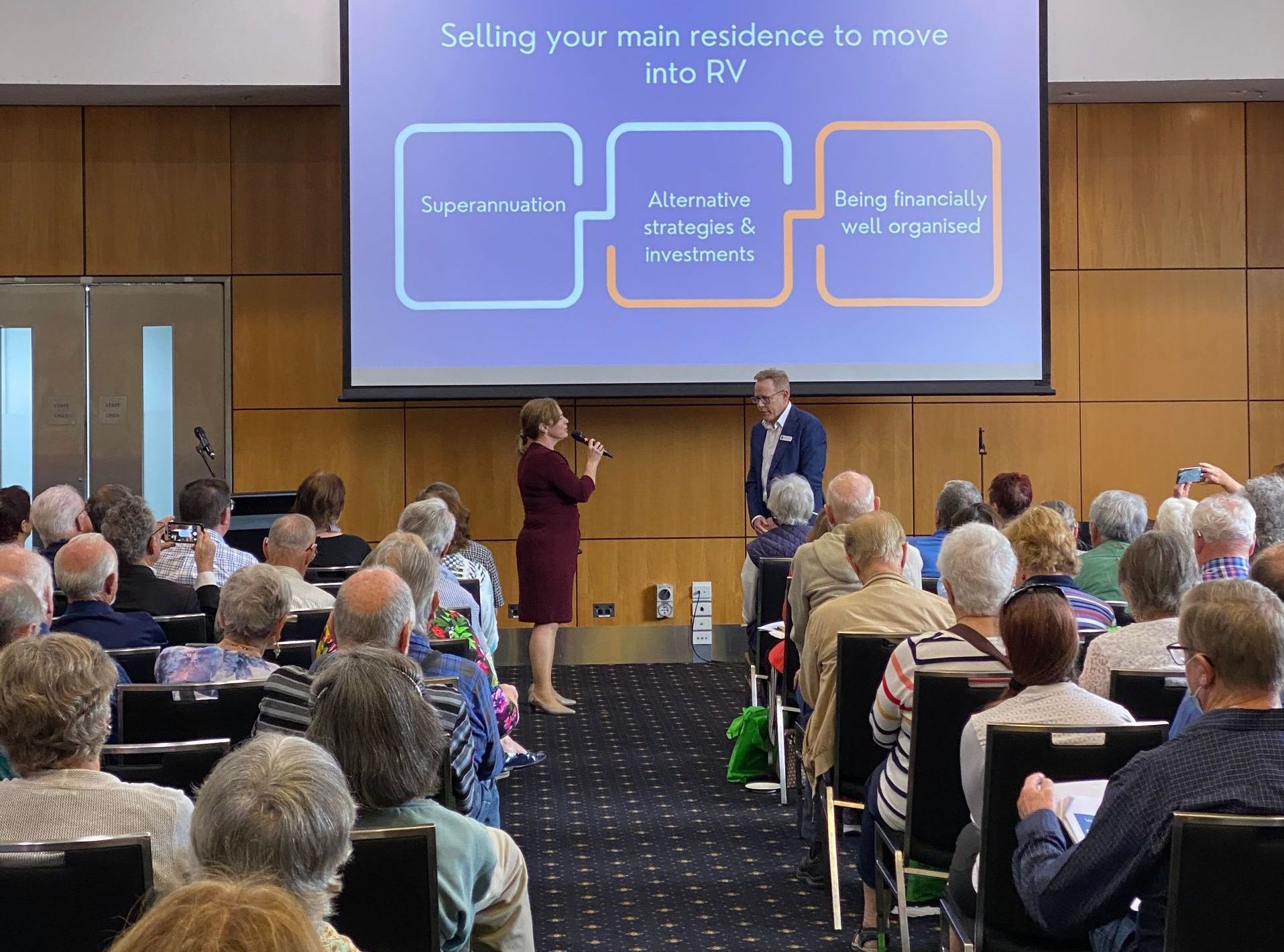 A presentation on selling a main residence to move into an RV. Two presenters, a projected graphic, and an audience in a large room.