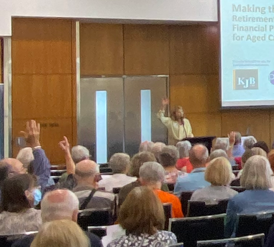 Woman lecturing at a retirement seminar. Attendees raise hands.