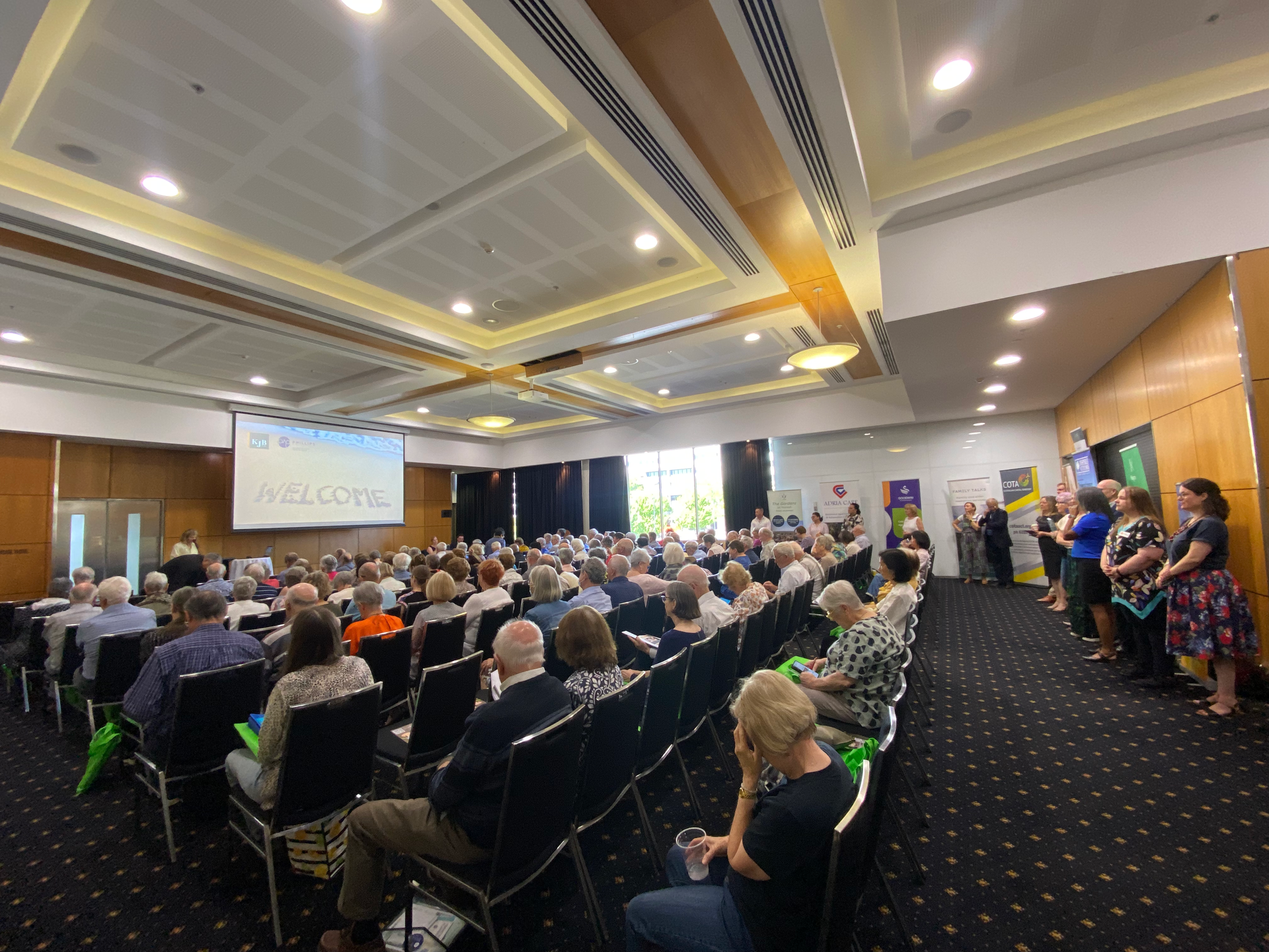 Large conference hall with seated audience, a welcome screen, and people standing on the side.