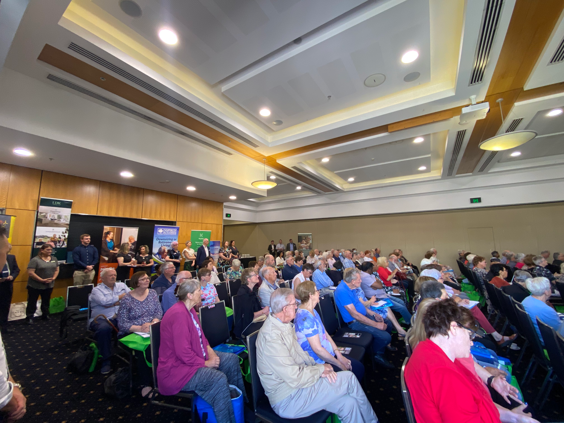 Audience seated in a large conference room, listening to a presentation; mostly older adults.