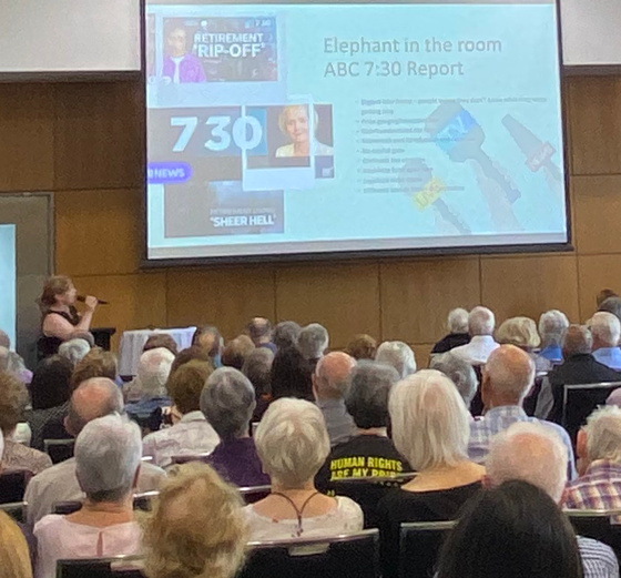 Woman speaks at event, screen shows ABC 7:30 Report graphics, audience seated, elderly people.