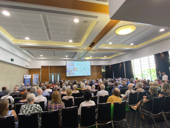 Large conference room filled with people attending a presentation. A screen displays information.