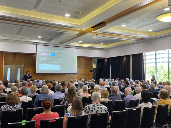A speaker presents to a large audience in a conference hall, with a projected screen showing information.