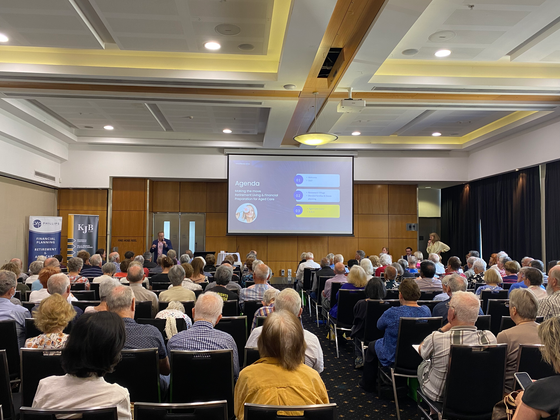 Audience at a conference presentation. A speaker stands at a podium, presenting a slide with text and graphics.