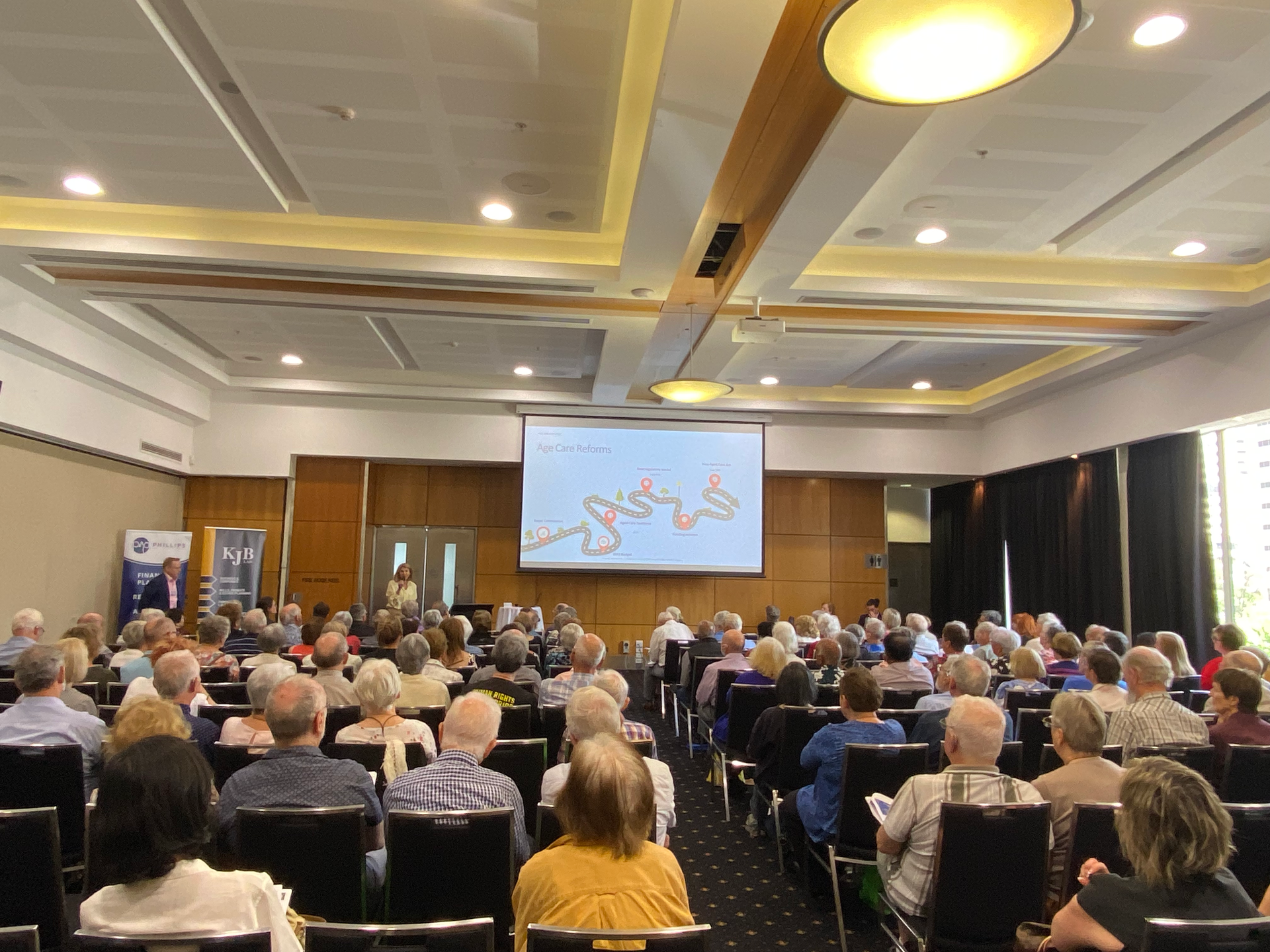 Audience in a large conference room, listening to a presentation on a screen.