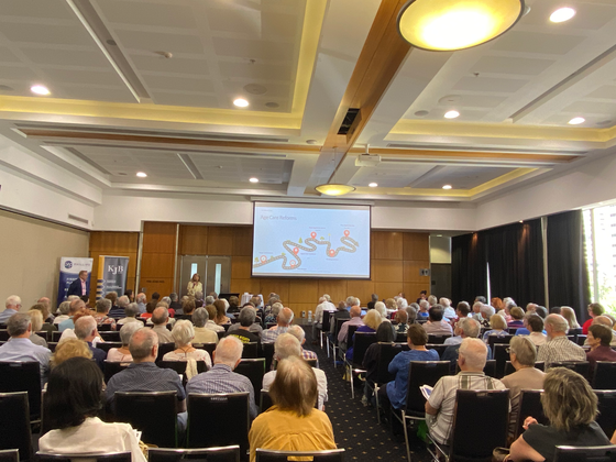 Audience in a large conference room, listening to a presentation on a screen.