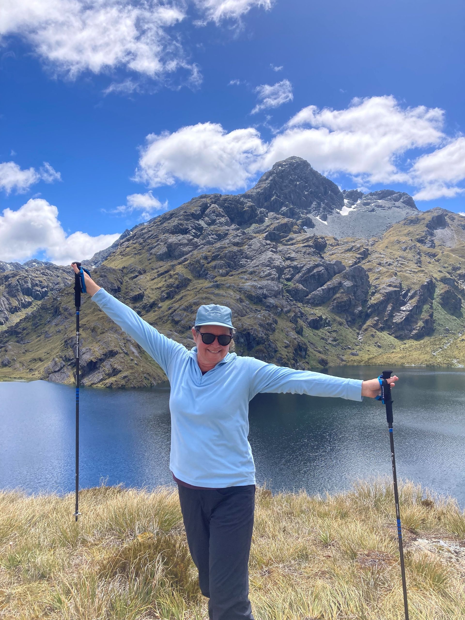 Woman with arms outstretched poses by a mountain lake. Blue sky, mountain backdrop.