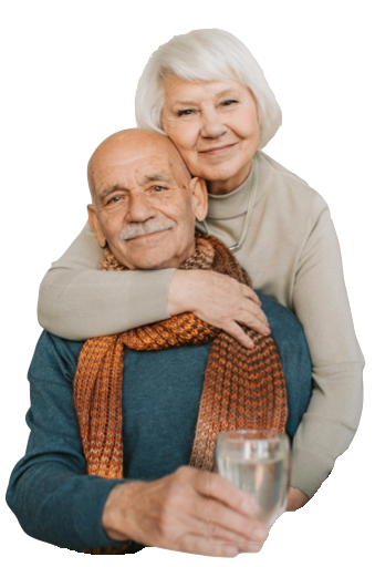 Senior couple embraces, the woman behind the man, smiling. The man holds a glass of water.