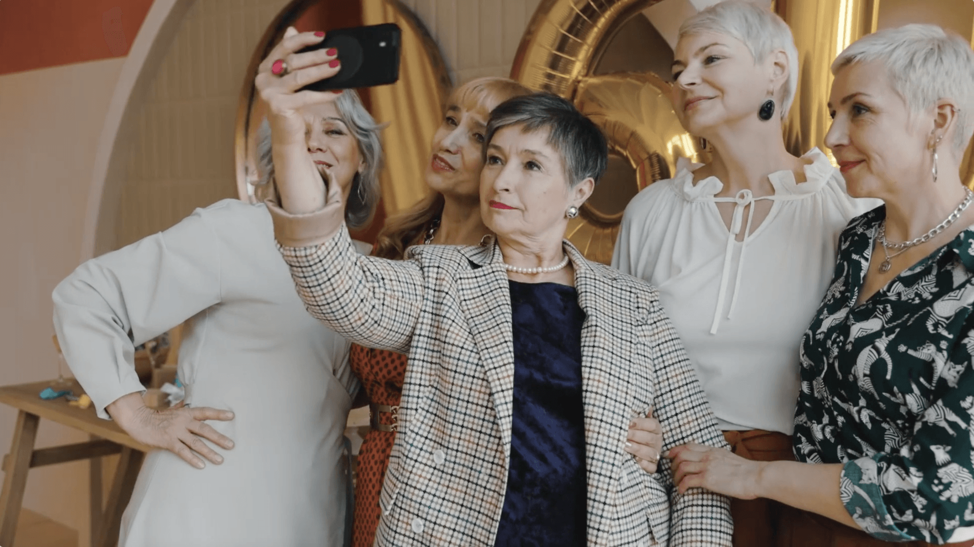 Five older women taking a selfie indoors; smiling, holding each other, and dressed in nice clothes.