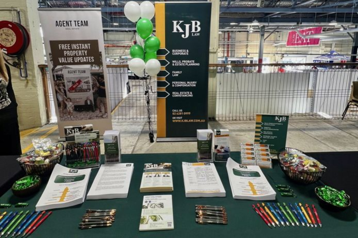 A display table at an event, with KJB branding, marketing materials, pens, and green/white balloons.