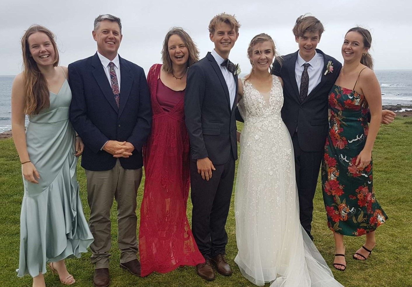 Family at wedding: bride & groom in formal wear, with parents and siblings on a grassy bluff overlooking the ocean.