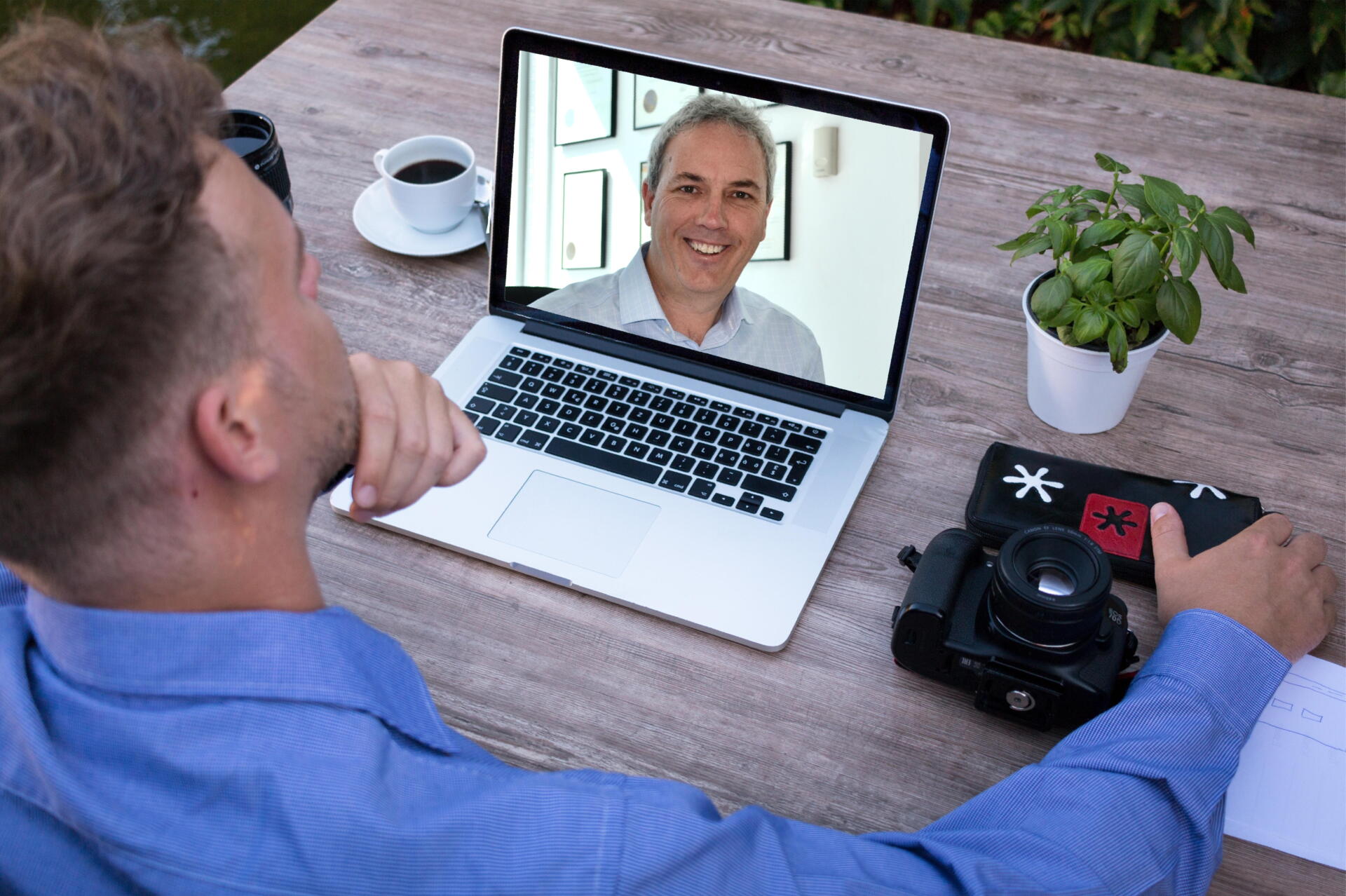 Man on video call with another man, laptop on table, coffee cup, camera.