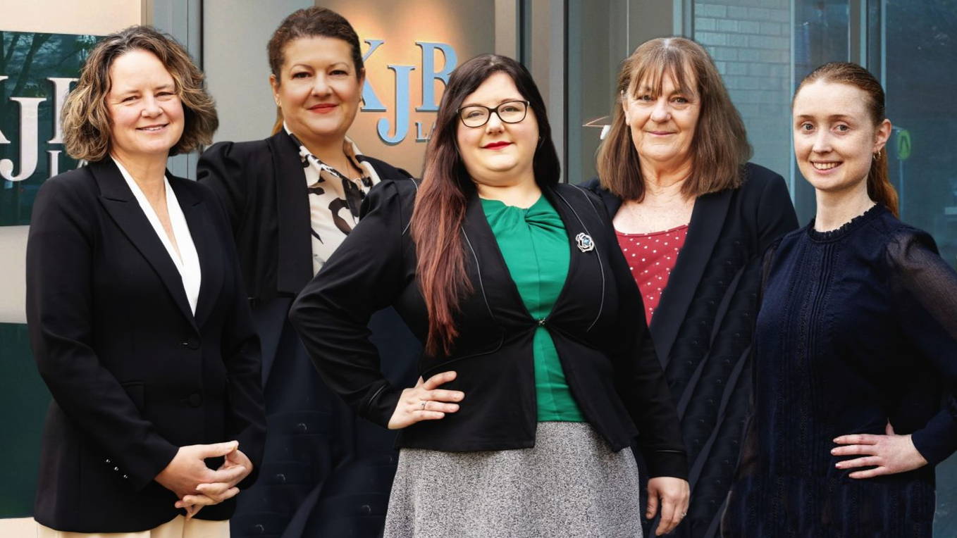 Five women in business attire pose outside a building with initials in the window.