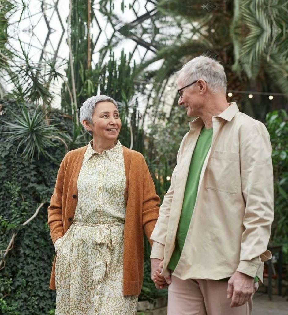 Elderly couple holding hands, walking together, smiling in a botanical garden.