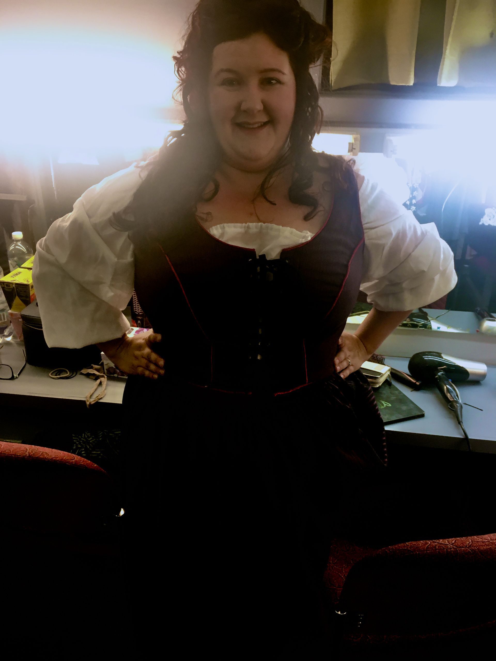 Woman in costume, arms on hips, smiling. Wearing white blouse, purple vest, and dark skirt. Inside a backstage area.