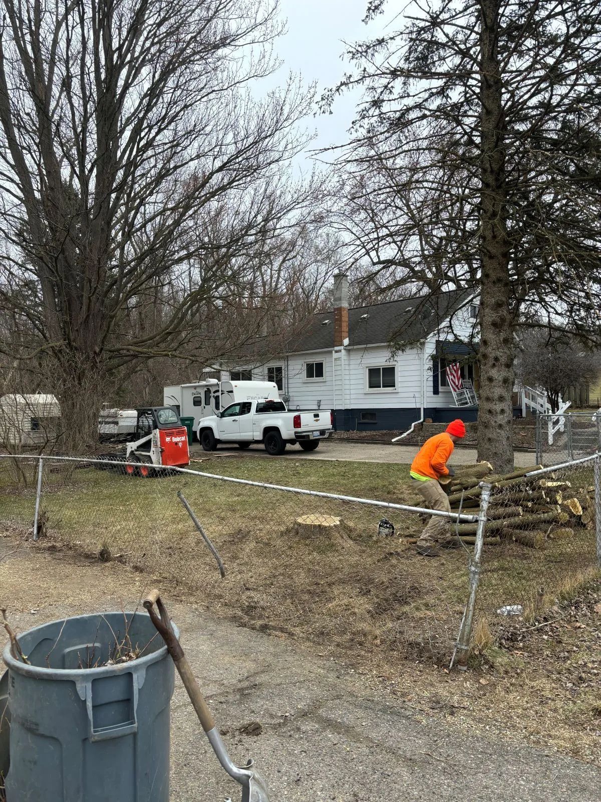 Man in orange shirt cutting wood near a white house with a truck parked in the driveway.