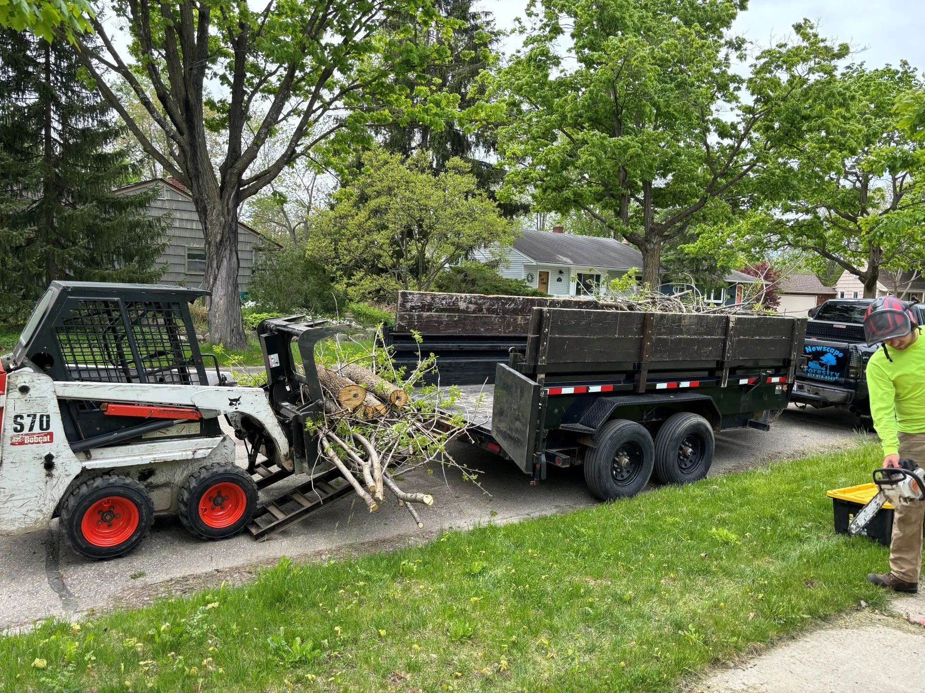 Bobcat loader loading debris into a trailer on a residential street; a person with a chainsaw stands nearby.
