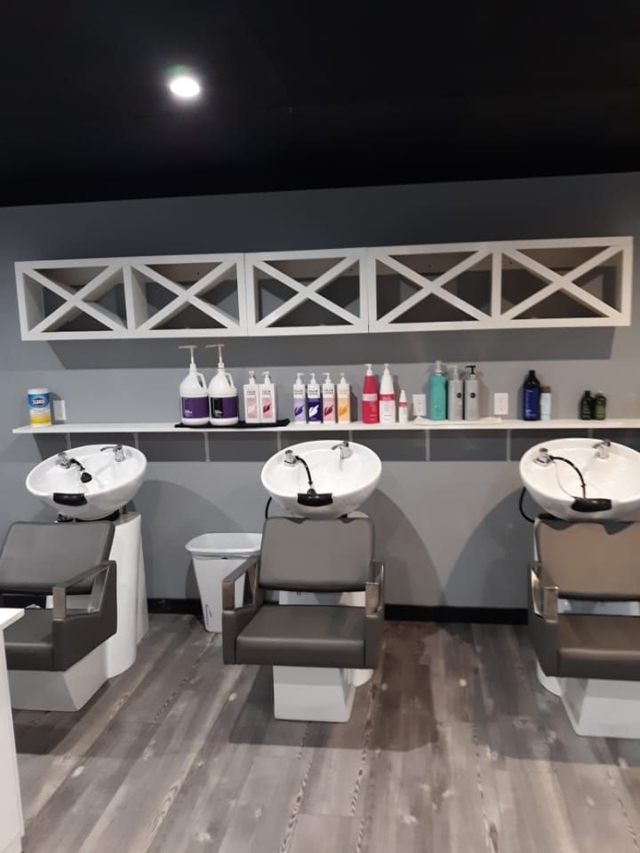 Empty salon wash station with three black shampoo chairs, white sinks, and product shelves on a gray wall.