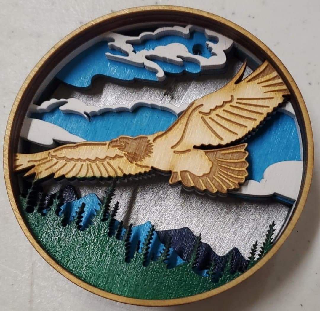 Wooden round wall art of an eagle flying over mountains, trees, and clouds in blue, tan, and green.