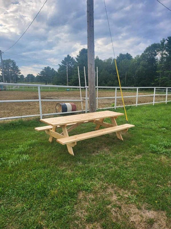 Wooden picnic table on grass beside a utility pole and white fence under a cloudy sky