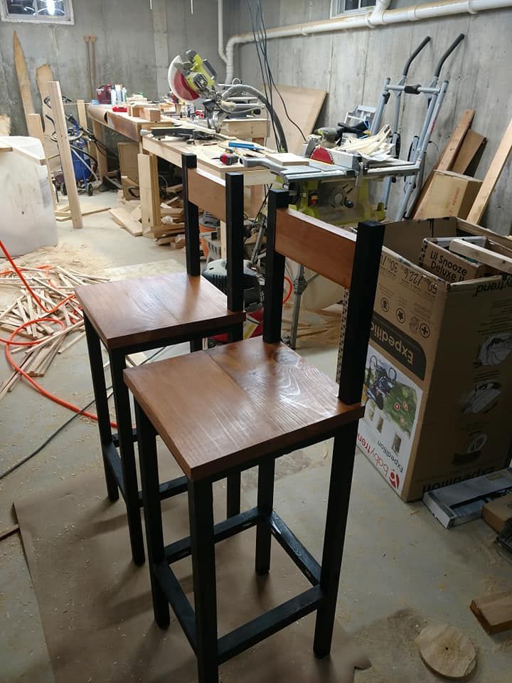 Two tall wooden stools in a cluttered workshop with workbench, tools, and boxes in the background