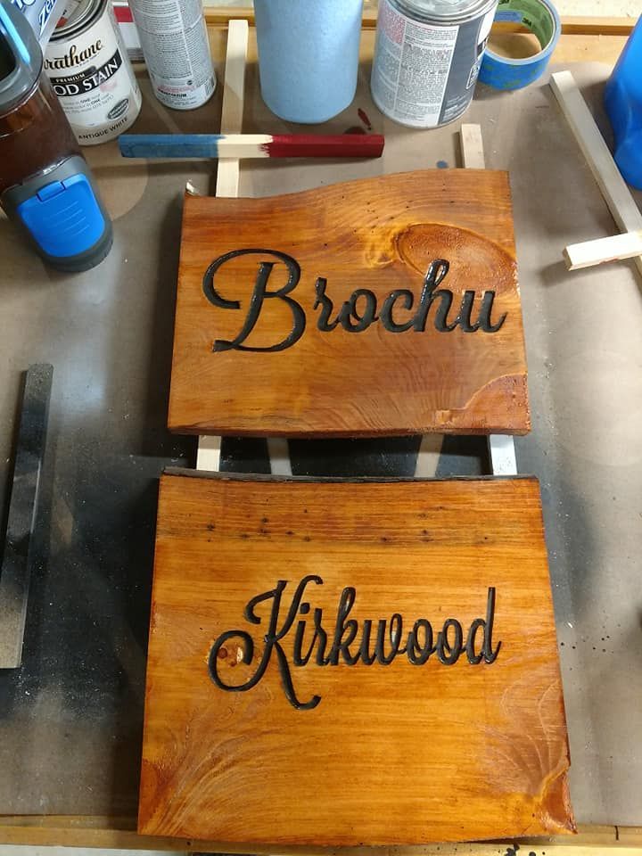 Two wooden name plaques on a workbench, stained brown and engraved with “Brochu” and “Kikwood.”