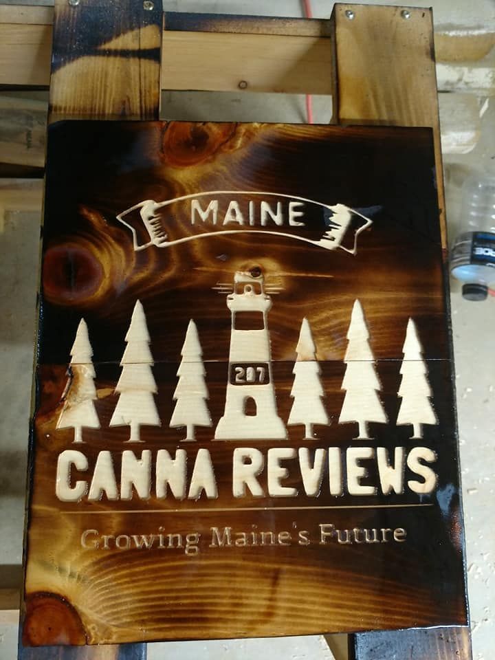 Maine Can-Am Reviews sign with lighthouse and pine trees on a dark wood background, “Growing Maine’s Future”