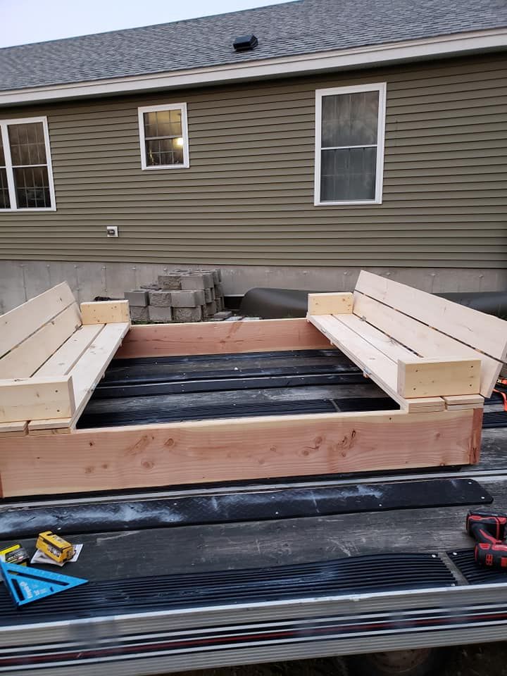 Wood-framed raised deck under construction beside a house, with stacked cinder blocks and tools on a trailer bed.