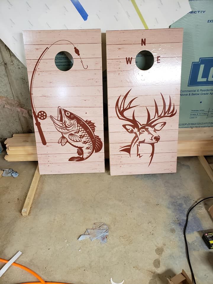 Two wooden cornhole boards with fish and deer designs, leaning against a wall in a workshop.