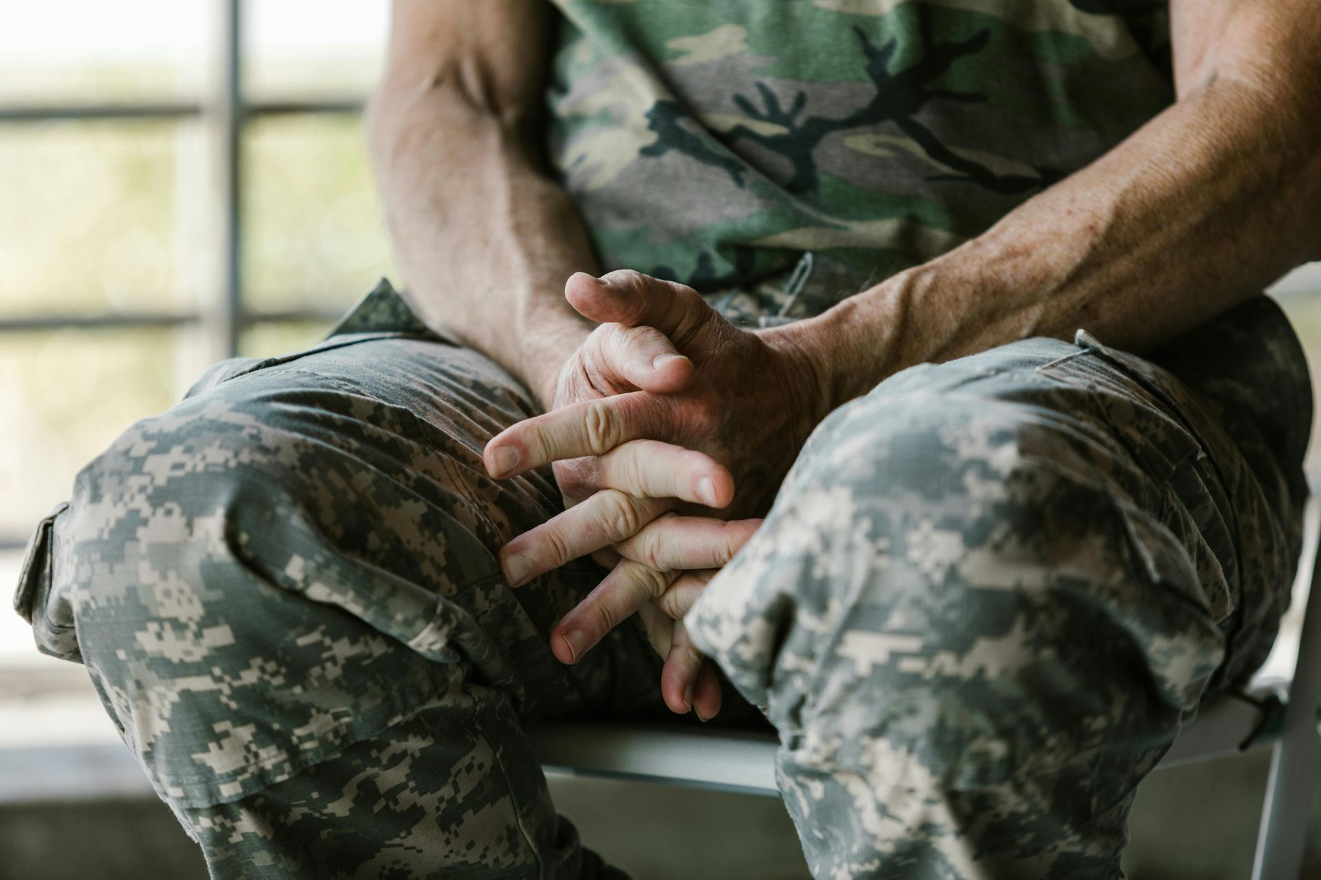 Person in camouflage, hands clasped, sitting, possibly in a waiting area.