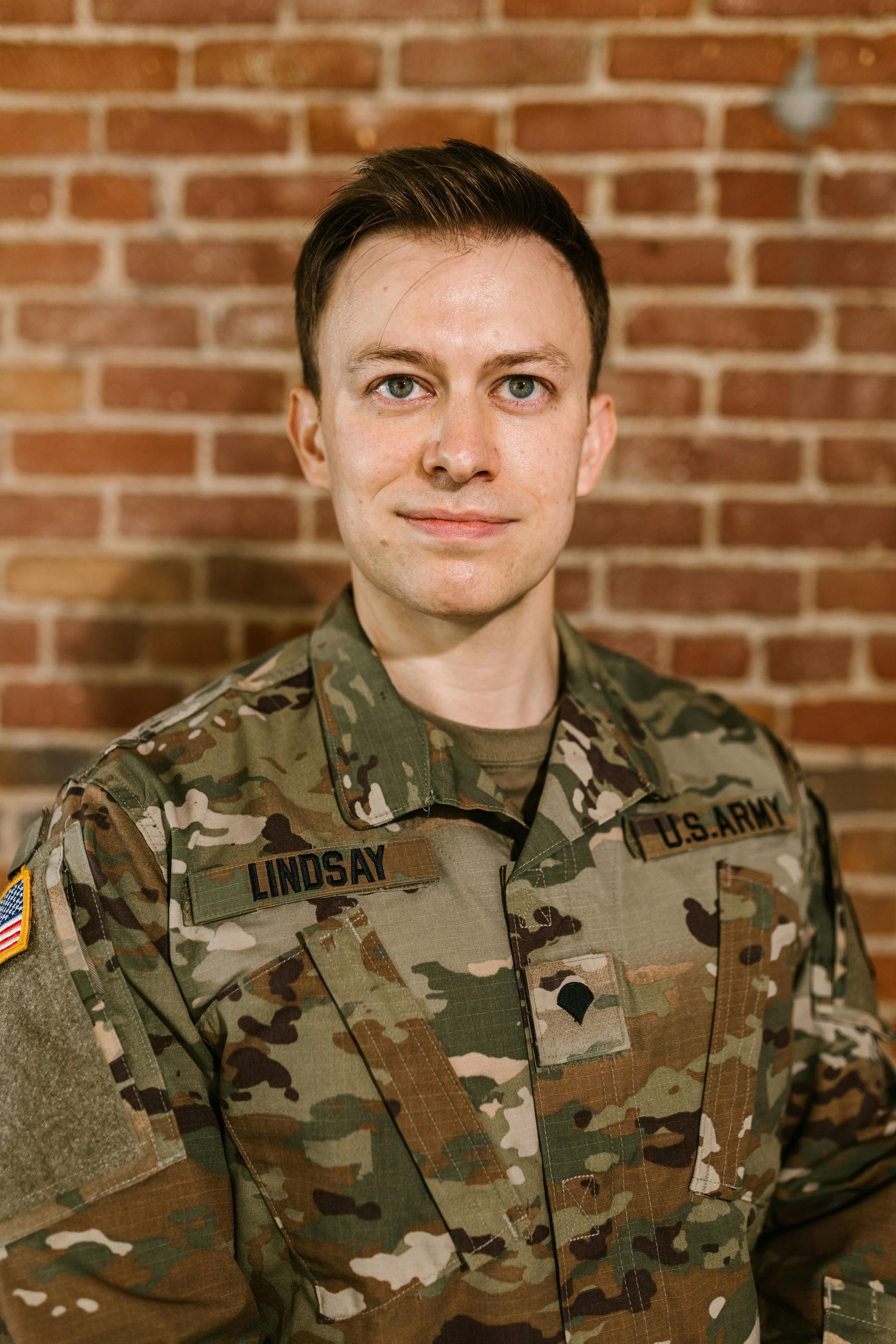 Man in U.S. Army uniform stands in front of a brick wall, smiling. Name tag: Lindsay.