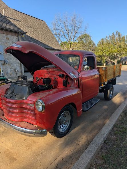 An old red truck with the hood up is parked on the side of the road.