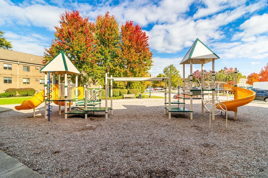 A playground with two structures featuring yellow slides, set against a backdrop of trees with autumn foliage.