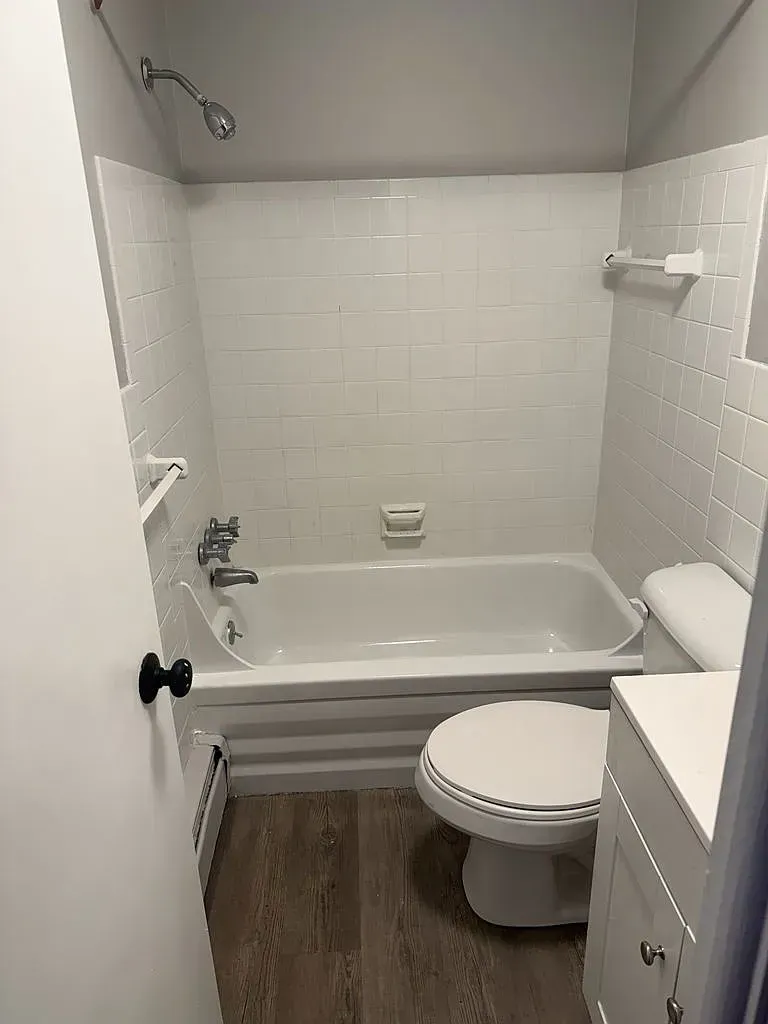 A small bathroom featuring a white bathtub, subway-tiled walls, a showerhead, a toilet, and light-toned wood-look flooring.