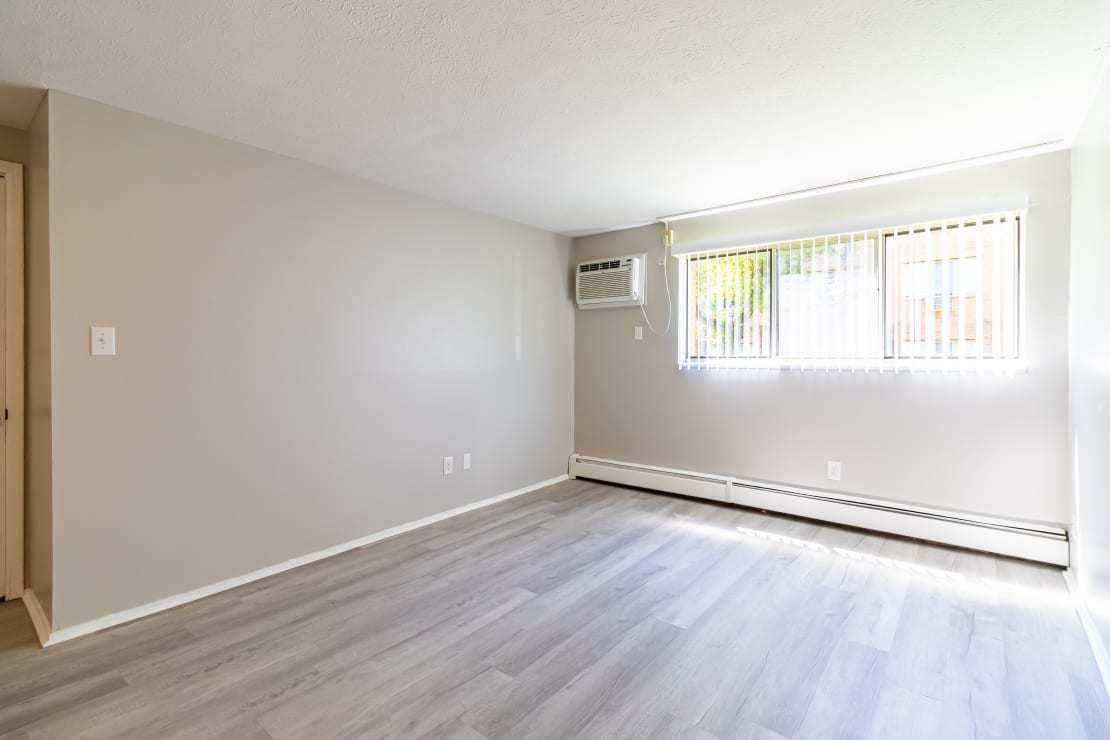 An empty, light-filled room with beige walls, light wood-style flooring, and a large window with an air conditioner unit.