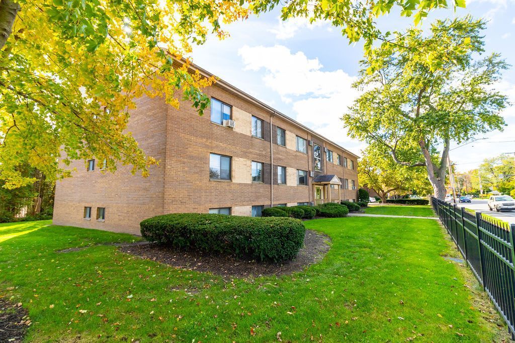 A three-story brick apartment building set on a lawn with autumn trees and a black metal fence on a sunny day.