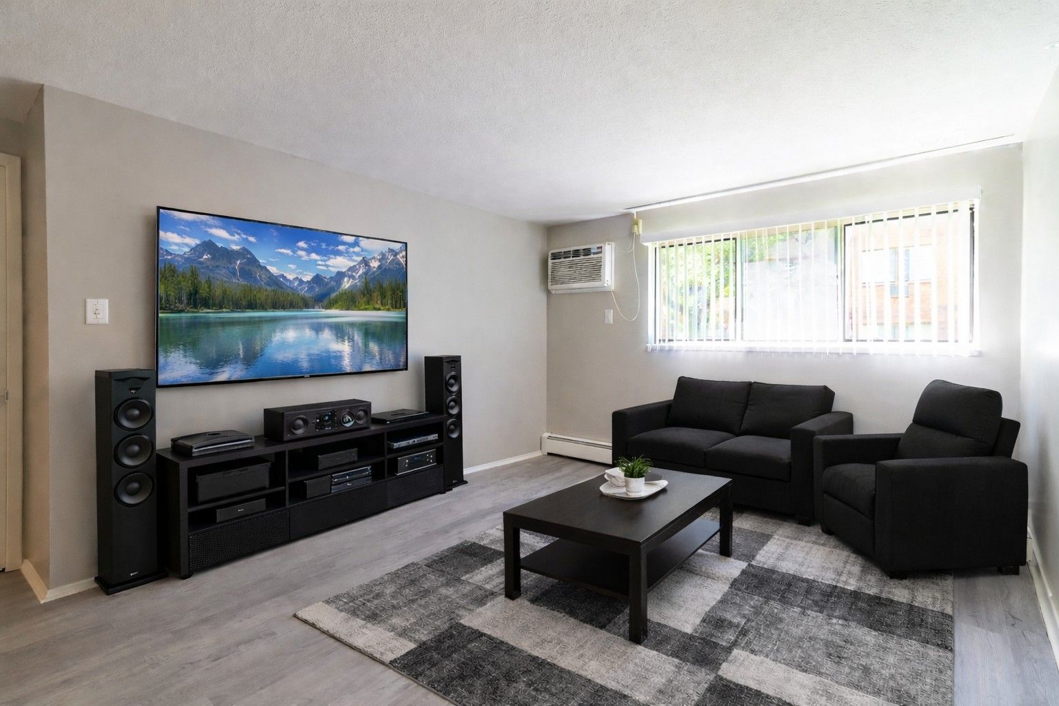A living room featuring a large wall-mounted TV, black speakers, media console, sofa, armchair, and a patterned rug.