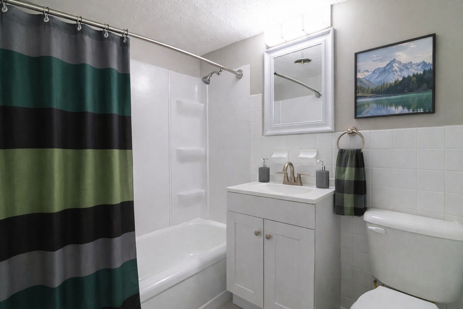 A clean bathroom with a striped shower curtain, white vanity, mirror, toilet, and a mountain scene framed on the wall.