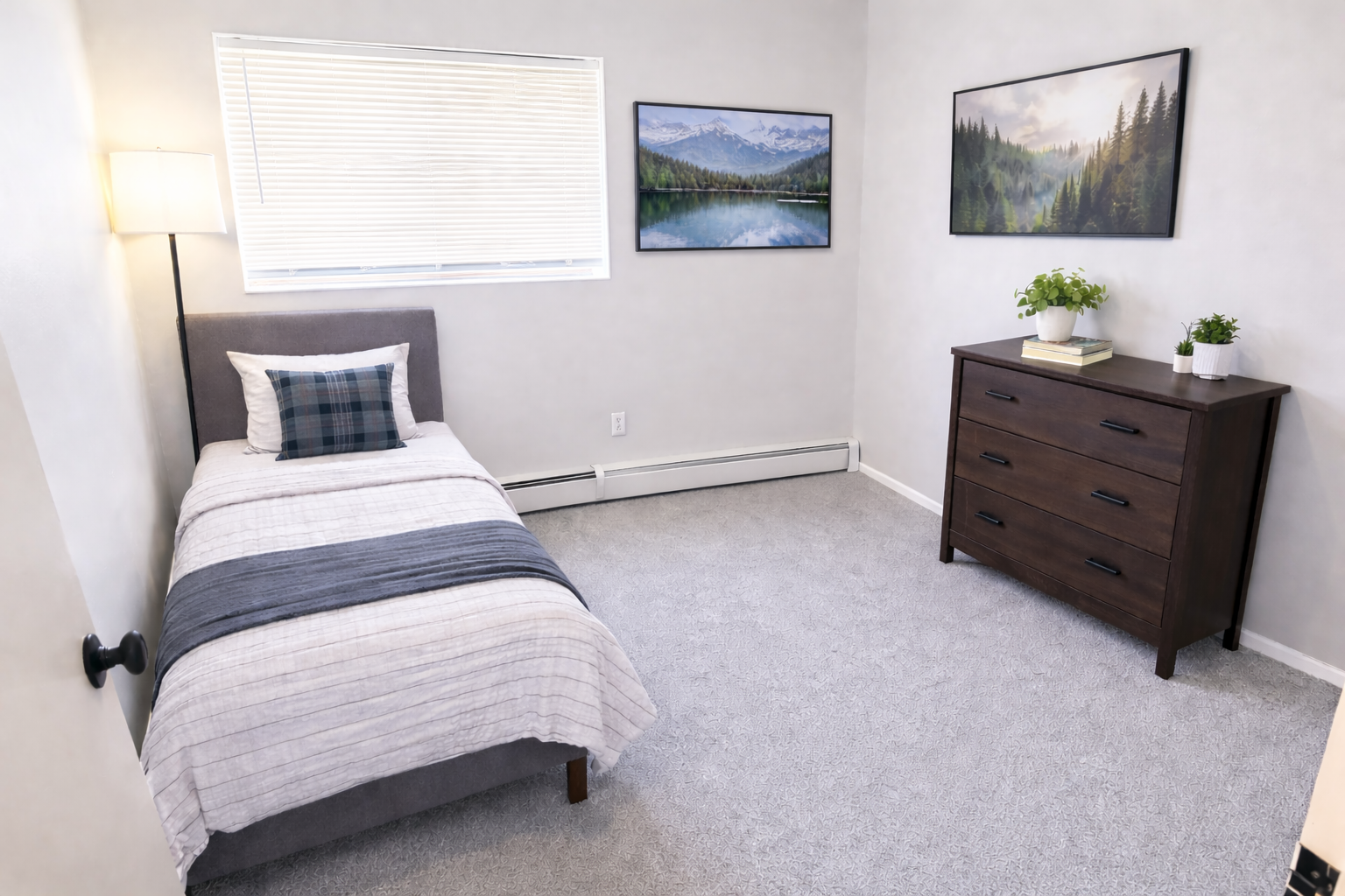 A furnished bedroom featuring a twin bed with grey bedding, a dark wood dresser, two landscape paintings, and wall lamps.