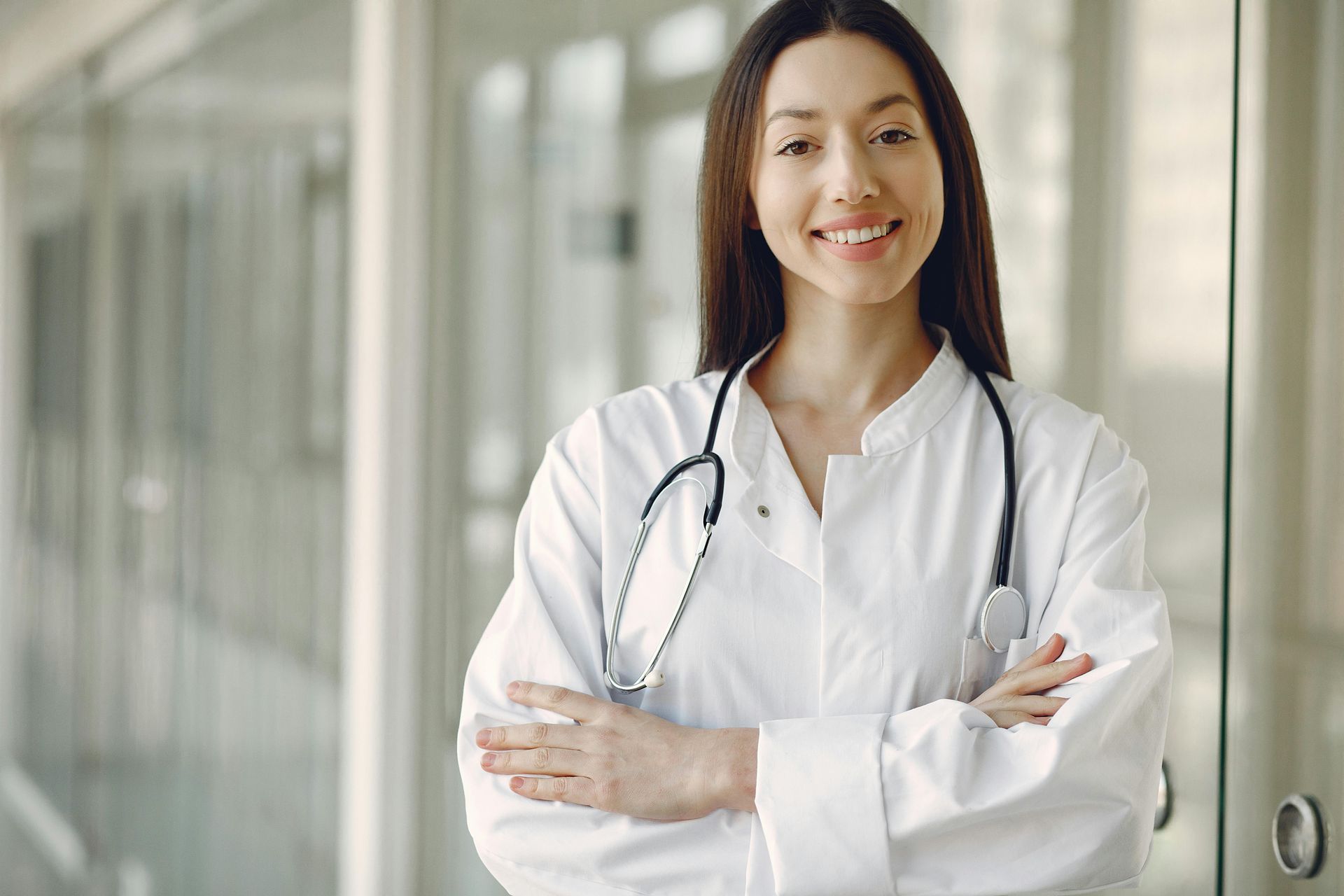 Woman in a white coat with a stethoscope around her neck, smiling with arms crossed in a brightly lit hallway.