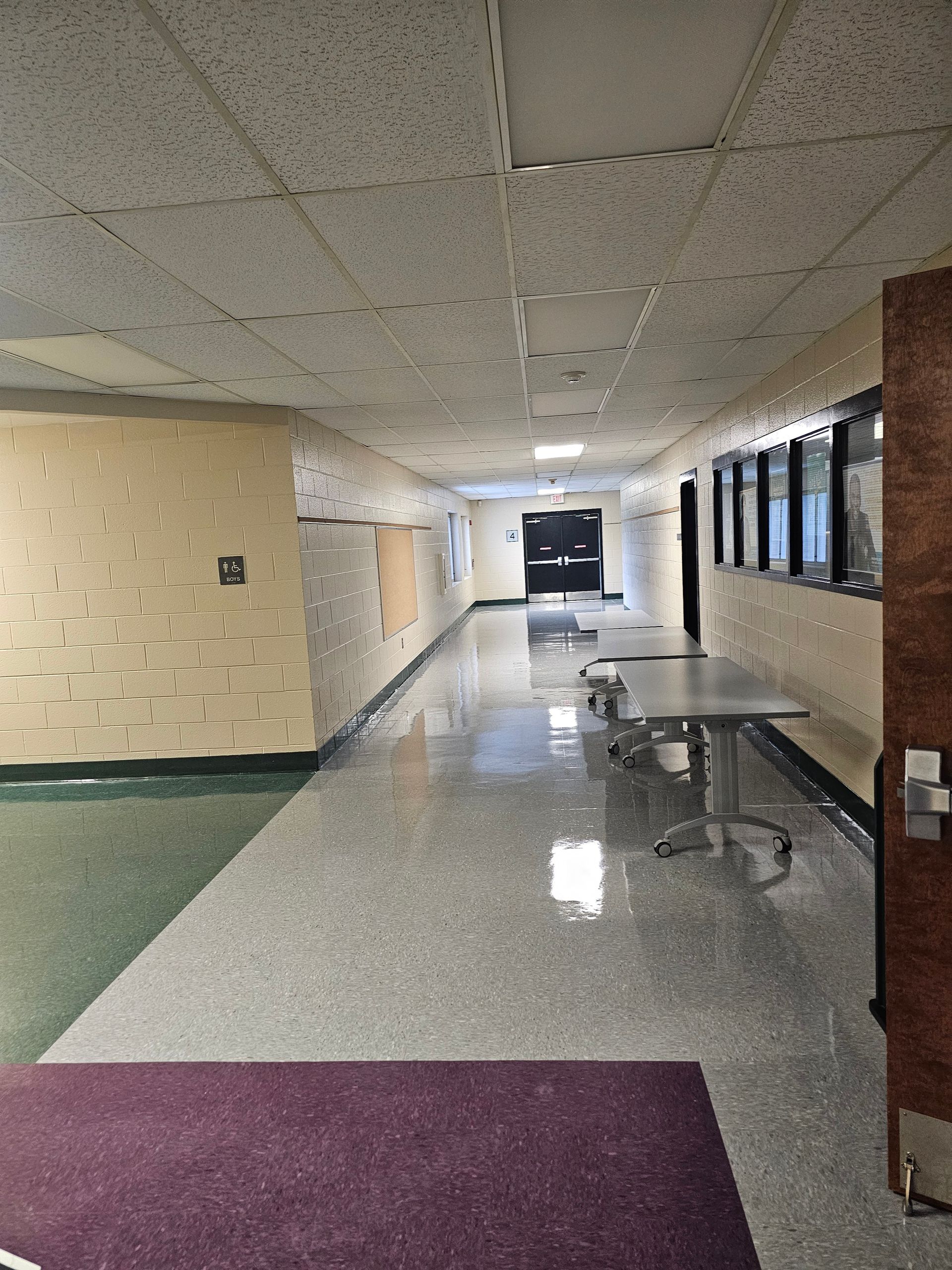 School hallway with tables, black door at the end, and a dark purple welcome mat.