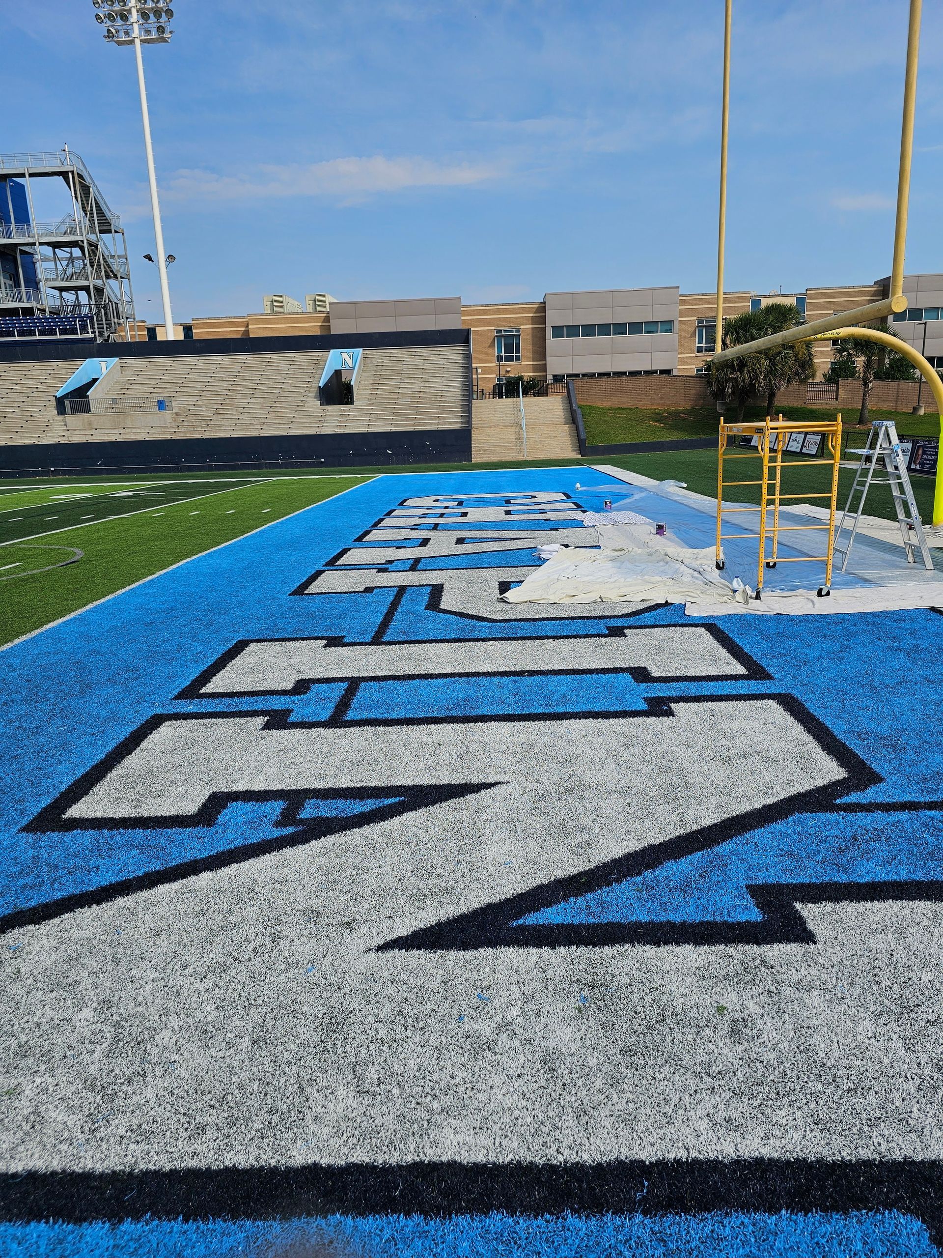 A football field with the school's logo painted in endzone, blue turf, bleachers in background.
