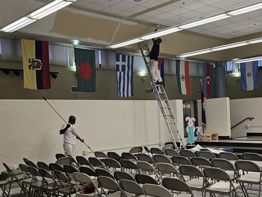 Two people painting a ceiling in a room with flags, rows of chairs, and a stage.