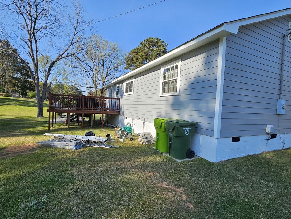 Side of a light gray house with a wooden deck, two green trash cans on grass under a clear blue sky.