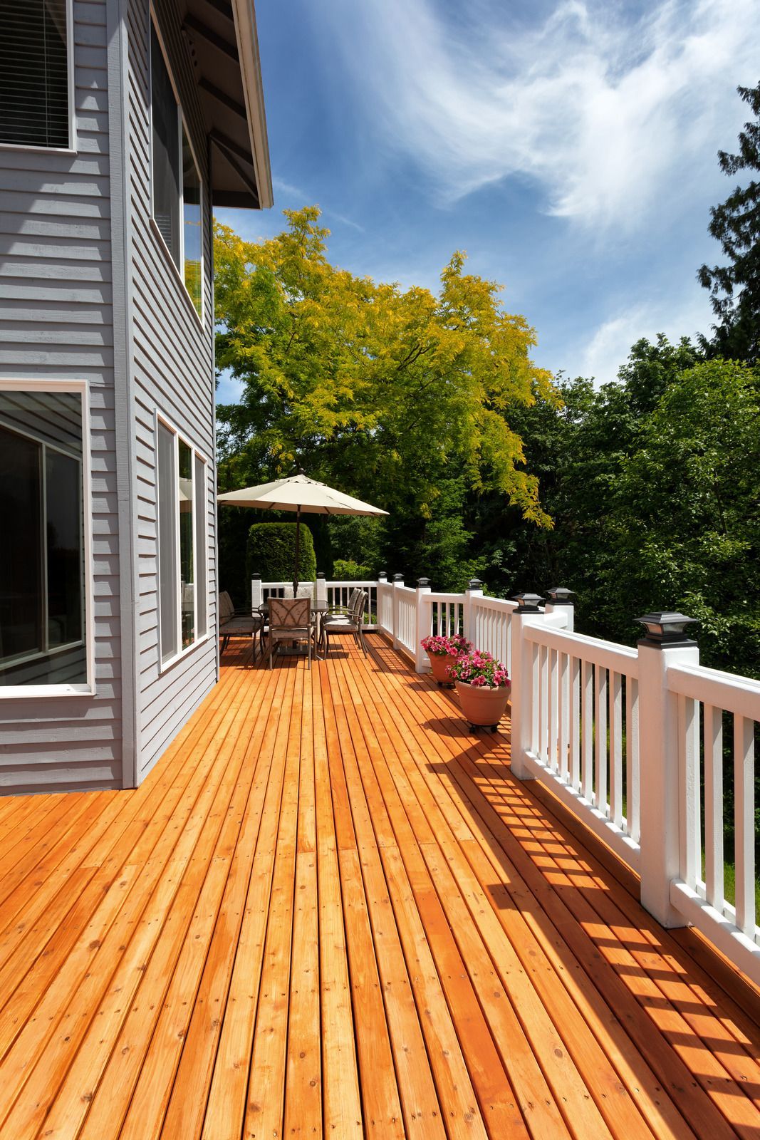 Wooden deck with white railing and patio furniture; gray house exterior; lush green trees, sunny sky.
