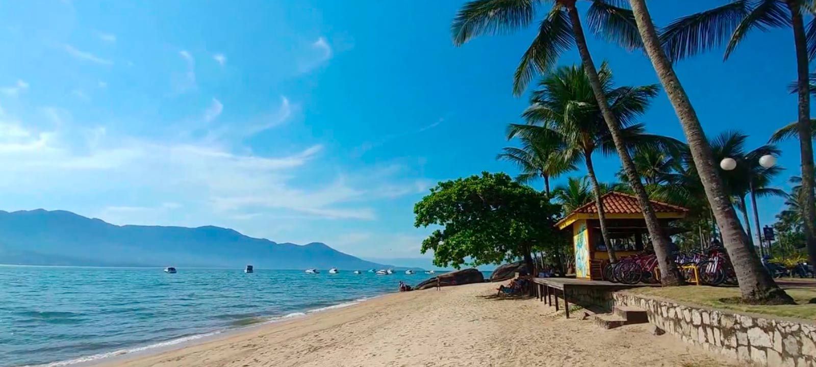 Uma praia tropical ensolarada com palmeiras, uma pequena cabana de madeira e montanhas distantes do outro lado da água.