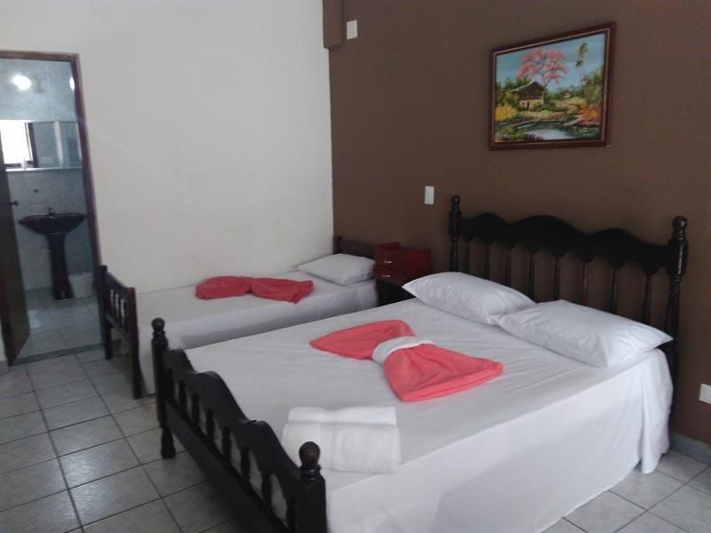 Um quarto com uma cama de casal e uma cama de solteiro com lençóis brancos, toalhas rosa e estrutura de madeira escura, próximo a um banheiro.
