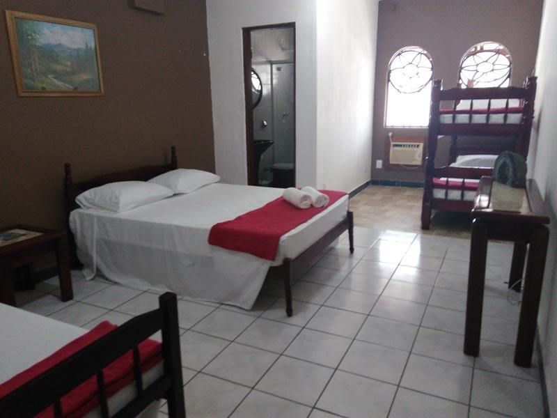 Um quarto de hotel com uma cama queen-size com um caminho de mesa vermelho, um beliche, uma mesinha de centro, uma porta para o banheiro e piso de azulejo.