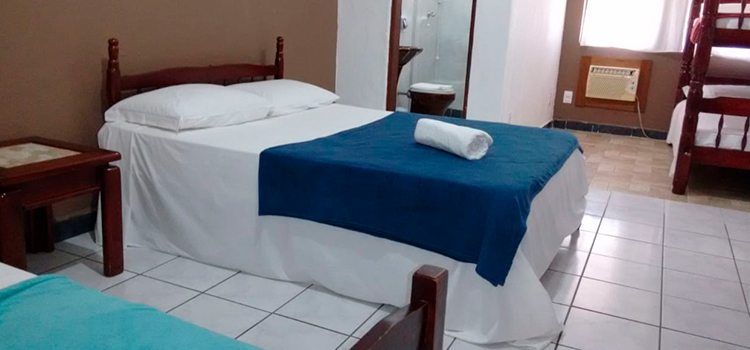 Um quarto de hotel com uma cama de casal com lençóis brancos e uma manta azul, uma mesa de cabeceira de madeira e um beliche no canto.