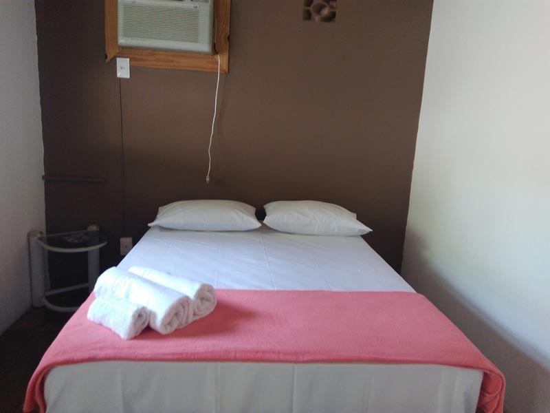 Uma cama impecavelmente arrumada com lençóis brancos e uma manta rosa, com toalhas brancas dobradas, encostada numa parede castanha do quarto.